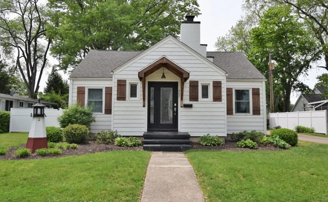 Blue cottage with yellow trim, stone foundation, and yellow front door on grassy lot with mature trees